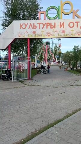 The video shows the entrance to a park in Russia. There are several small motorized vehicles parked near the gate. The sign above the entrance says "Park Kultury i Otdykha" which translates to "Park of Culture and Recreation."  A Ferris wheel can be seen inside the park. A few people are visible in the park.  The ground is paved with grey bricks. There are trees in and around the park, and the sky is a mix of blue and white.  The gate to the park is made of metal and painted red and white.  A price list is attached to the fence to the left of the gate.