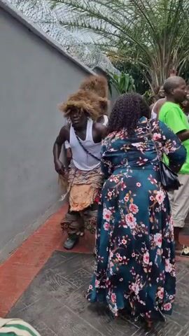 A group of men in traditional attire with large, furry headdresses are performing a dance. One man in the foreground, wearing a white tank top, patterned skirt, and leg adornments, is bent over as he dances. A woman in a floral dress is standing behind him, appearing to interact with him. Other men in similar attire and a man in a green shirt are visible in the background. The scene takes place outdoors next to a grey wall with a wire fence on top, surrounded by vegetation.