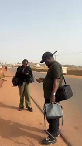 Two people, a woman and a man, stand on the side of a road. The woman is wearing a black jacket and green pants, and has a backpack. She holds a phone in her hand and is facing the camera and making a peace sign with her fingers. The man is wearing a green shirt, black pants, and a black baseball cap. He has two bags slung over his shoulders and is looking down at a phone in his hand. The road is paved and has a red dirt shoulder. There are buildings and power lines in the background. The sky is pale blue.