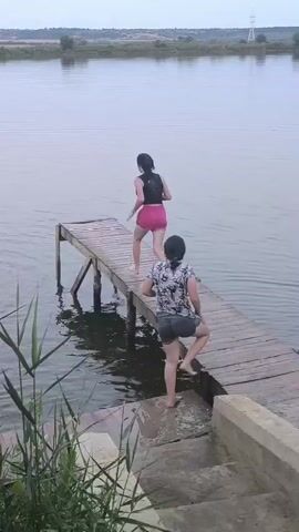 Two young women are on a wooden pier extending into a body of water. The woman closer to the shore is wearing a patterned top and shorts and is walking towards the end of the pier. The woman further down the pier is wearing a black top and pink shorts and runs towards the edge. She jumps into the water, creating a splash. The second woman then also jumps into the water. Reeds are visible in the foreground on the left side of the frame, and a distant shoreline with trees and buildings can be seen. The sky is overcast.