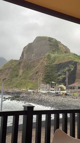 The video shows a view from what appears to be an enclosed patio or porch, looking out at a rocky coastline. The ocean is in the foreground, with waves crashing against the shore. Beyond the shore are mountains covered in green vegetation. The sky is overcast and foggy, obscuring the peaks of the mountains. There's some infrastructure visible along the coastline, including a street, some buildings, utility poles, and what may be construction equipment. A black wooden railing runs along the bottom of the frame. People are heard speaking in the background. The camera slowly pans from right to left.