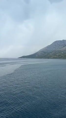 The video captures a sweeping view of a body of water, likely the sea, under a cloudy sky. The water is a deep blue, with gentle ripples visible on the surface. In the distance, a mountainous coastline is visible, characterized by its rocky slopes and sparse vegetation. The sky is overcast with heavy, gray clouds, suggesting an impending storm or the aftermath of one. The video pans across the scene, offering a panoramic perspective of the expansive water and the rugged landscape. There are no people or man-made objects explicitly shown in the frame, focusing solely on the natural environment. The overall atmosphere is one of vastness and a somewhat somber, yet majestic, natural beauty.
