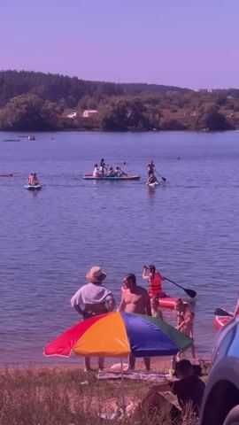 A video shows a sunny day at a lake. In the foreground, several people are gathered on the shore. A colorful umbrella is set up. Some individuals are sitting or standing near the water's edge. In the mid-ground and background, numerous people are on the lake, many of them on paddleboards. Some groups are on larger paddleboards, appearing to be recreational outings. The lake is surrounded by trees and a gentle slope of land. The water is calm, and the sky is a clear, light purple hue, suggesting a bright and warm day.