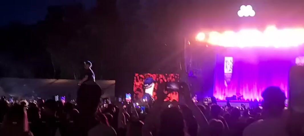 The video shows a nighttime outdoor concert. A large crowd of people is gathered, many holding up their phones. A stage is visible with bright purple and orange lights. On the stage, a performer is singing into a microphone. A large screen behind the performer displays a close-up of their face. In the foreground, a person is being carried on someone else's shoulders.
