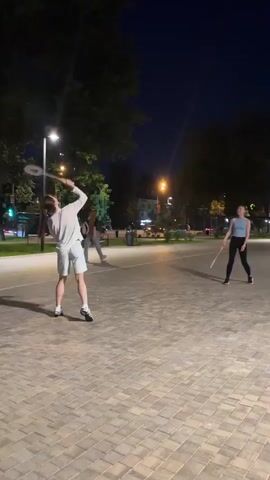 Two women are playing badminton in a park at night. The woman on the left is wearing a white t-shirt and light blue shorts, and is in the process of hitting a shuttlecock. The woman on the right is wearing a light blue tank top and black leggings, and is preparing to hit the shuttlecock. There are streetlights and trees in the background, and the ground is paved with interlocking stones.