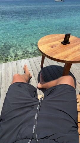 The video shows the first-person perspective of someone sitting in a chair on a wooden deck overlooking the ocean. The person is wearing black shorts, and their bare feet are visible. Next to the chair is a small, round wooden table with a table number stand that reads "4". The ocean is a clear, turquoise color, and there are rocks visible through the water near the deck. In the distance, there's a white motorboat and some land visible on the horizon, and some white puffy clouds dot the blue sky.