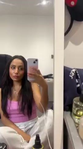 A woman sits on the floor, facing a mirror. She is holding a purple iPhone with a colorful pop-it case and taking a selfie. She is wearing a pink tank top and white sweatpants. Her hair is long and dark. The background shows a plain white wall, a black leather chair, and a side table with various objects on it. A white framed mirror reflects the scene.