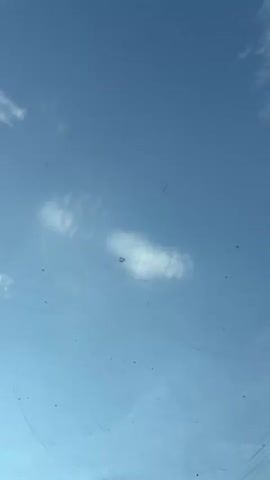 The video shows a view from inside a vehicle, looking up through a sunroof and out a side window. The sunroof reveals a clear blue sky with a few wispy clouds and some small dark specks that could be insects or dust. The side window offers a panoramic view of a lush, green mountainous landscape with a body of water visible in the distance. In the foreground of the side window, there is a white railing. The interior of the vehicle is visible at the bottom, with a dark fabric headliner, a grab handle, and what appears to be an air freshener hanging from it. The overall impression is of a scenic drive.