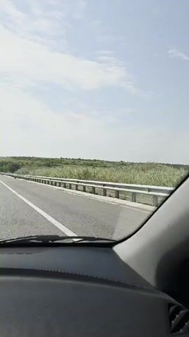 This is a first-person perspective video taken from inside a car driving on a highway. The dashboard is visible in the foreground. The highway has multiple lanes and is bordered by a guardrail and green hills with some trees. The sky is partly cloudy. Other vehicles can be seen on the road ahead. The video is short, lasting only a few seconds.