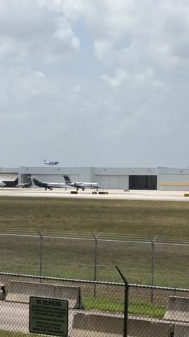 A medium-shot video shows an airport scene with multiple aircraft and hangars under a cloudy sky. In the foreground, a chain-link fence with barbed wire runs horizontally across the bottom of the frame. Behind the fence, a grassy area is visible, followed by a concrete barrier. A white sign is attached to the concrete barrier, reading "NO TRESPASSING." In the mid-ground, two private jets are on the tarmac. The jet on the left is white with a dark stripe and appears to be moving or stationary. The jet on the right is also white and appears to be moving. In the background, large white hangars with dark entrances are visible. Further in the distance, a light blue airplane is in the air, ascending or descending against the sky. The overall scene depicts a typical airport operational area.