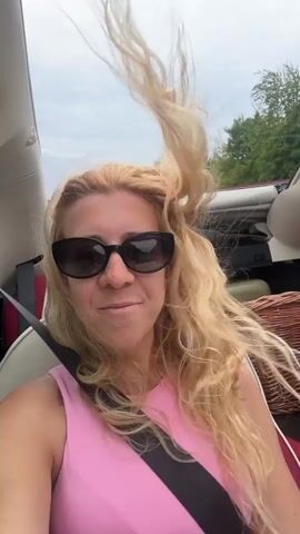 A woman with blonde hair and sunglasses, wearing a pink top, is sitting in a car with her hair blowing in the wind. She smiles at the camera and raises her right arm. There is a seatbelt across her chest. In the background, there are trees and a cloudy sky.