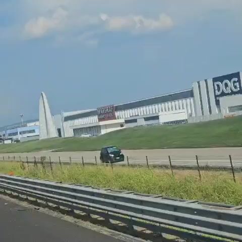 The video shows a large industrial building with the "MARGRAF" logo visible on the facade. A distinctive white arched structure stands in front of the building. A black car is driving on a road in front of the building. The environment appears to be a commercial or industrial area with a blue sky and white clouds. The camera is moving, suggesting it is being recorded from a moving vehicle.