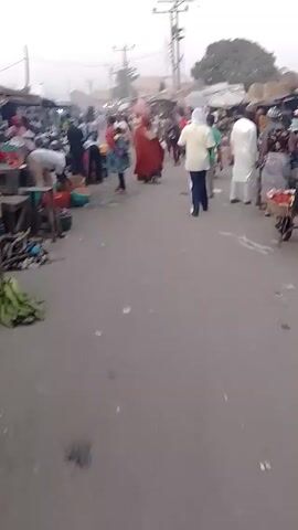 The video depicts a crowded outdoor market, likely in an African setting, given the attire of many of the individuals. Numerous people walk along a paved road lined with vendors and their wares. The market appears to be selling produce, with vegetables and fruits visible in colorful containers. There is a moderate amount of litter on the ground. The overall atmosphere seems busy and lively. The sky is overcast, creating a slightly hazy or muted light.