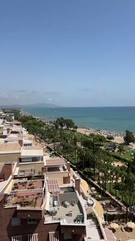 A drone view pans right over a coastal town with buildings in the foreground and a beach and ocean in the background. The sky is clear and blue. The buildings are multi-story structures with balconies and rooftop patios. Palm trees line a road between the buildings and the beach. The beach is dotted with umbrellas and beachgoers. The ocean is a calm blue.
