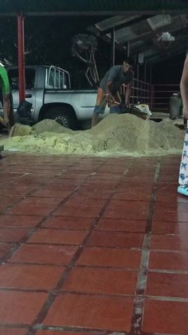 Two men are shoveling sand under a covered area with red-tiled flooring. A silver pickup truck is parked behind them. The men are wearing casual clothes; one has on a green shirt and the other a gray shirt and orange shorts. A child in pajamas is partially visible on the right side of the frame. The setting appears to be outside at night, with artificial lighting illuminating the scene.