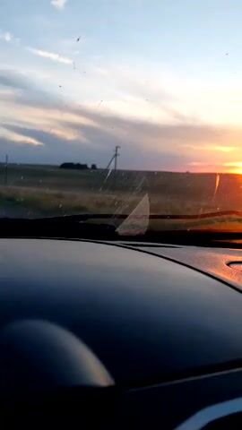 The video shows a sunset viewed from inside a vehicle. The sun is low on the horizon, casting an orange glow across the sky. Fields and a utility pole are visible in the distance. The foreground of the image is dominated by the dark, reflective dashboard of the car, with streaks on the windshield. The overall impression is of a peaceful, driving-through-the-countryside scene at dusk.