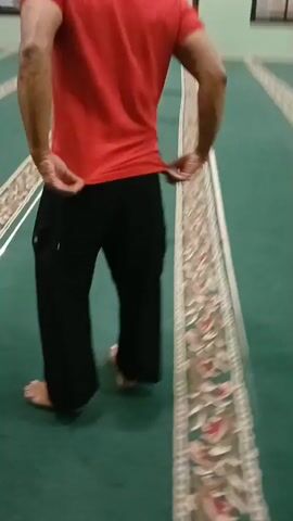 The video shows a group of people in a mosque. The camera pans across a green carpeted floor with decorative borders. Several men and boys are visible, some standing in lines, seemingly preparing for prayer. One individual in a red shirt and black pants walks across the carpet. The environment appears to be an indoor space with patterned walls and some decorative elements, including a circular emblem on one wall and a whiteboard or notice board on another. The lighting suggests it is daytime or well-lit artificial lighting. The overall atmosphere appears to be one of communal gathering or religious observance.