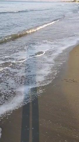 The video shows a beach with waves gently rolling onto the shore. The water is a clear blue, and the sand is a light brown. The sun is shining, casting a long shadow of the person filming onto the sand. There is no one else visible in the video, and no objects other than the natural elements of the beach. The video appears to be taken from a low angle, close to the water's edge.