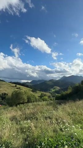 The video shows a panoramic view of a rural landscape with rolling hills, green fields, and scattered trees. The sky is a vibrant blue with fluffy white clouds. In the foreground, tall grass and small white flowers are visible. In the middle ground, the hills are covered in lush green vegetation, with a few trees and some buildings scattered among them. In the background, more distant hills and mountains are visible, partially obscured by haze. The scene suggests a peaceful and natural setting, possibly a countryside or a mountain region. There are no people or animals visible in the video. The lighting indicates it is daytime, likely late morning or early afternoon, given the bright sunlight and the shadows cast by the clouds.