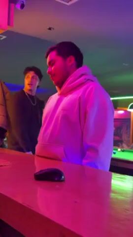 A young man in a light pink hoodie stands at a bar in a dimly lit room. He picks up a toothpick from the counter, puts it behind his ear, and smiles to himself. Another person is visible in the background, standing further away near a pool table. The room's lighting creates pink and green hues on the scene. A computer mouse sits on the bar counter in front of the man. A security camera is visible mounted on the ceiling.