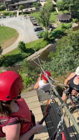 The video shows an aerial view of people zip-lining. In the foreground, two individuals wearing helmets and safety harnesses are preparing to zip-line. One person, wearing a red helmet and a red vest, is in the process of being hooked up to the zip-line. The other person, with a white helmet, is already attached and appears to be assisting or waiting. In the mid-ground, a third person, wearing a red helmet and a white shirt, is actively zip-lining across the scene, suspended from a cable. Below the zip-line, a river winds through a village with buildings, a parking lot, and a grassy area. The surrounding landscape is characterized by rolling green hills and mature trees. The weather appears to be sunny and clear.