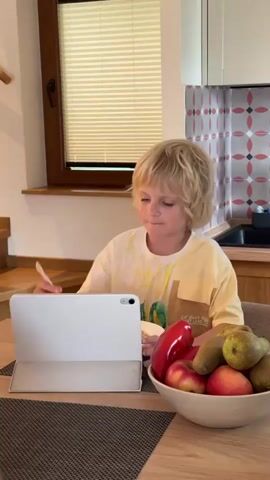 A young boy with blond hair is sitting at a wooden table and eating from a bowl with a fork while watching a tablet computer. The tablet is open and propped up on a white case. In front of the tablet is a bowl of fruit containing red apples, red bell peppers, and pears. The boy is wearing a light yellow t-shirt with a graphic design. The background shows a window with blinds, a wooden shelf, and a kitchen counter with cabinets. The lighting appears to be natural, coming from the window.