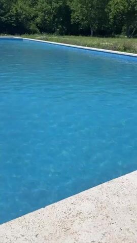 This video shows a close-up view of a swimming pool filled with bright blue water. The pool has a white concrete edge visible in the foreground, and a blue tiled border around the top edge of the pool. In the background, there are green trees and a hint of blue sky. The water is clear, and the sunlight creates rippling patterns on its surface. There are no people or other objects visible in the water. The camera slowly pans across the pool, providing a sense of its vastness and tranquility.
