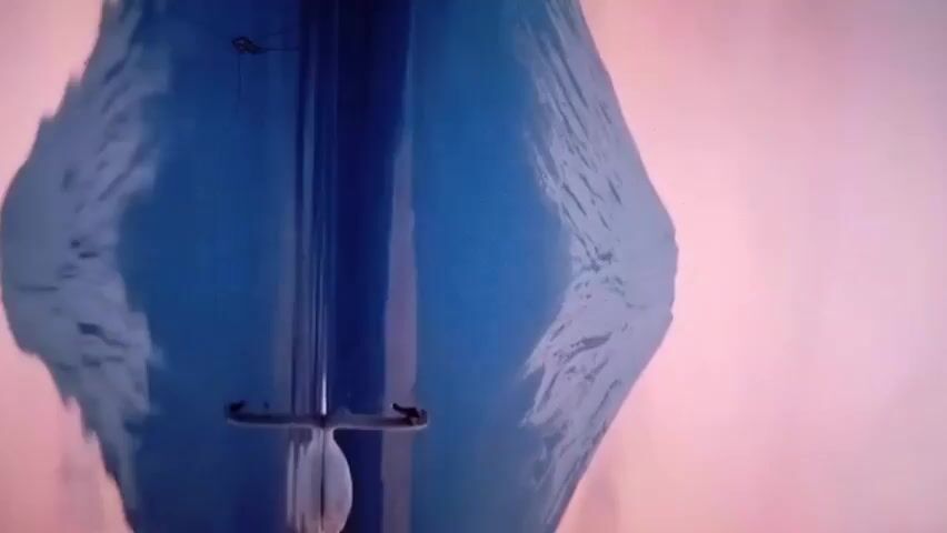 A close-up shot shows a portion of a wind instrument, possibly a cello or double bass, against a muted background that is light pink to white. The instrument's body is a rich, deep blue. A tuning peg is visible, and a small, white object hangs from it. The image is somewhat abstract due to the proximity of the camera to the subject.