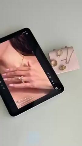A slightly angled overhead shot shows a white iPad displaying a vertically oriented social media video, alongside a small, light pink jewelry pouch and several gold rings and necklaces. The iPad screen showcases a woman wearing a thin gold necklace and an ornate ring with prominent stones on her hand, which is placed on her chest. The video then switches to a full-body shot of the same woman in a black spaghetti-strap dress. The jewelry pouch has a loop on top and contains a ring and necklace. The table's surface is a glossy, reflective white.