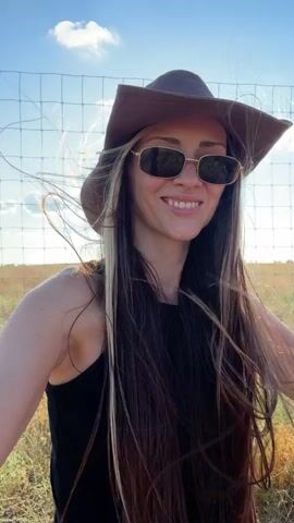 A woman wearing a brown cowboy hat and sunglasses smiles at the camera. Her long brown hair blows in the wind. She is wearing a black tank top and stands in a grassy field with a fence in the background. Beyond the fence, several brown animals, possibly bison, graze in the field. The sky is blue with a few white clouds.