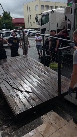 A group of men are gathered around a wooden platform that appears to be suspended by a crane. The platform is wet, suggesting it might have rained or been washed. Several men in the foreground and background are looking at the platform and the crane. One man, wearing a black t-shirt and shorts, is walking away from the platform. Another man, wearing a grey t-shirt, is standing near the platform and looking at it. In the background, a white truck is visible, as well as other parked cars and buildings. The sky is overcast.