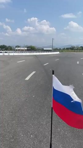 The video shows a Russian flag stuck in the ground by the side of an empty, asphalt road on a sunny day. The road has white lane markings. There is a white barricade to the left side of the road with blue arrows pointing to the left. In the background is a flat, green landscape and blue sky with puffy clouds.