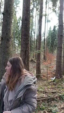 The video shows a person standing in a wooded area, looking down a slope covered with trees and ground vegetation. The individual is wearing a tan puffer jacket and a light pink hoodie. They are holding a phone in their left hand. The forest consists mainly of tall, thin trees, with some deciduous trees mixed in. The ground is covered with fallen leaves, moss, and some patches of snow. A glimpse of a village or town is visible in the distance through the trees, partially obscured by the forest density. A red mark can be seen on one of the trees close to the camera.