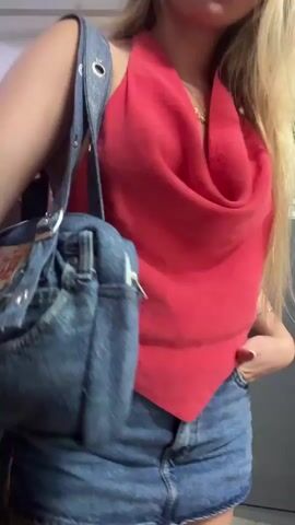A young, slender, fair-skinned woman with long, straight blonde hair stands in a parking garage, posing for the camera. She is wearing a red backless halter top with a cowl neck, a denim miniskirt, and a denim shoulder bag with a silver chain. She has a gold necklace and a silver bracelet on her left wrist. Her fingernails are painted white. Her left hand is in her skirt pocket, and her right hand holds the strap of her bag. She begins by facing the camera, then turns to her right, showing her left profile, then turns to her left, showing her right profile, then turns back to face the camera. She smiles faintly and appears confident. The background is a white wall with a red fire extinguisher cabinet and a gray car on the left. On the right is a gray door with an exit sign.
