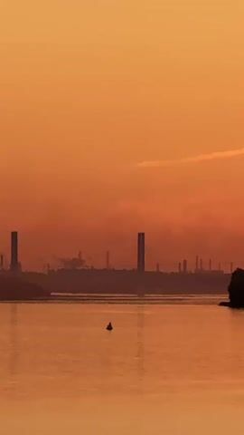 The video shows a wide shot of a body of water with an industrial landscape in the distance. The sky is a vibrant orange, indicating either sunrise or sunset. In the foreground, the water reflects the warm colors of the sky, creating a shimmering effect. There is a small, dark object, possibly a buoy or a marker, in the water. The middle ground features a silhouetted shoreline with trees and buildings, including tall, rectangular structures that appear to be industrial towers. Smoke or steam is visible emanating from some of these structures. Further in the distance, a dark, forested area lines the horizon. The video pans slightly to the right, revealing more of the industrial skyline and the tree line. A single bird is seen flying across the orange sky. The overall impression is one of an industrial setting at dawn or dusk, with the natural beauty of the sky and water providing a striking contrast to the man-made structures.