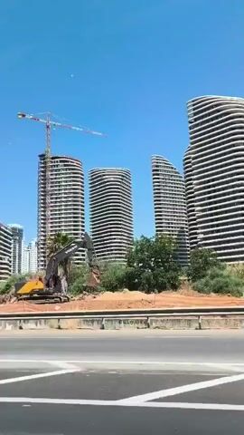 The video shows a construction site with several tall, modern buildings under construction. A large yellow excavator is parked on the dirt ground in front of the buildings. A tall crane stands behind one of the buildings. In the foreground, there is a road with a concrete barrier. The sky is clear and blue. The buildings have a unique curved design with horizontal white lines. Some trees and bushes are visible around the construction site. A white car is partially visible on the left side of the screen in the foreground.