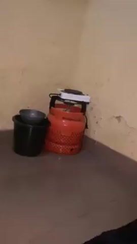A dimly lit corner shows a tan wall with peeling paint. On the floor against the wall are a black plastic bucket containing a silver bowl, an orange gas canister with a black net covering, and a small white rectangular electrical appliance set on top of the canister. The floor is dark brown and tiled. A small window is visible high on the wall.