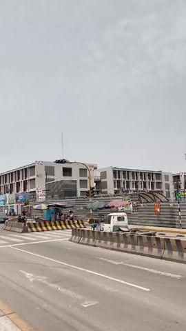 The video shows a street scene on a cloudy day.  There are several pedestrians walking along the sidewalk and crossing the street.  Cars are driving on the road.  In the background, there is a large, modern-looking building with red accents. There are barriers and construction equipment visible in the median of the street.  A traffic light is also visible, showing a green light.  There is a sign on a pole with a phone number on it.