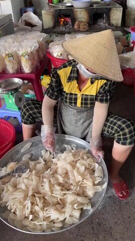 A person, likely a woman, wearing a traditional Vietnamese conical hat (nón lá), a face mask, a yellow and black plaid shirt, plaid pants, an apron, arm sleeves, and clear gloves, is actively mixing ingredients in a large metal basin. She is squatting or kneeling, using a spoon and her gloved hands to stir a large quantity of white, flaky pieces, which appear to be coated with a white, possibly sugary, substance.

The setting is an indoor or semi-outdoor food preparation area in Thới Sơn, Vietnam. In the background, a traditional wood-fired stove with visible flames indicates cooking or heating processes are underway. Various items associated with food production, such as baskets, plastic bags containing packaged goods (suggesting finished products), and other kitchen utensils, are scattered around the space. A green weighing scale is visible on the left side of the frame. The overall atmosphere suggests a small-scale, traditional food production operation.