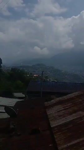A panoramic view of a hilly, urban landscape unfolds under a cloudy sky. The foreground is dominated by the rusty corrugated metal roofs of several buildings, one with a satellite dish positioned on it. Beyond these structures, the mid-ground reveals a dense cluster of buildings spread across the sloping terrain, suggesting a town or city nestled within the hills. The background consists of hazy, mist-covered mountains and hills, indicating a potentially humid or rainy atmosphere. The overall impression is one of a densely populated area situated in a mountainous region.
