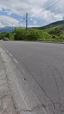 The video shows a view of a road in Bazum, Armenia, from a sidewalk. The road is asphalt, with a concrete curb and white dashed lines marking the lanes. Several vehicles are traveling on the road. In the foreground, a white truck with a blue tarp covering its cargo bed is moving away from the camera. Further down the road, a black car is passing the truck. In the distance, a brown sedan is visible on the road, and beyond that, a white SUV and a dark SUV are driving. The scene is set in a rural or semi-rural area with rolling green hills in the background. Trees and vegetation line the side of the road. Utility poles with power lines are also visible. The sky is partly cloudy with white clouds against a blue sky, suggesting it is daytime with fair weather. The overall impression is a snapshot of everyday life on a road in a scenic, mountainous region. There is no visible text in the video.