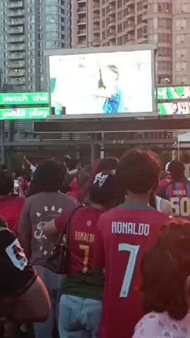 The video shows a crowd of people gathered outdoors watching a large screen displaying a soccer game. Many people in the crowd are wearing soccer jerseys, including several with the name "RONALDO" and the number 7. The screen shows a close-up of two male soccer players interacting on the field. Behind the screen, there are tall buildings and a stage set up for an event, with lights visible. The overall atmosphere appears to be one of a public viewing event for a sporting match.