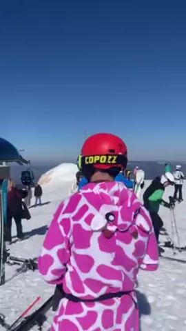 The video shows a busy ski resort on a sunny day. People are gathered around an outdoor restaurant, sitting at tables and enjoying the atmosphere. There's a ski lift nearby, and many skis are piled up in the snow. One person is wearing a pink and white cow-print jumpsuit and a ski helmet. The sky is clear blue.
