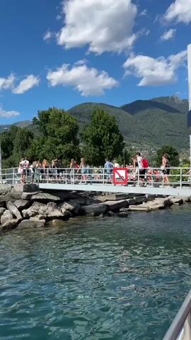 The video shows a group of people standing on a pier with a mountainous landscape in the background. The pier has a metal railing and a rocky edge. A red and white sign is visible on the railing. Some people are walking along the pier, while others are standing and looking out at the water. The water is blue and calm, with ripples on the surface. The sky is blue with white clouds. The overall scene appears to be a scenic view of a lake or body of water on a sunny day.