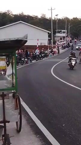 The video shows a street scene with a curve in the road. On the left side of the road, there is a building and a row of parked motorcycles. Several people are standing near the motorcycles. On the right side of the road, there are more parked motorcycles, food vendors with carts and umbrellas, and people gathered around. Cars and motorcycles are driving on the road. The environment appears to be an outdoor setting, possibly near a coastal area, with some trees and foliage visible in the background. The sky is overcast or it is dusk.