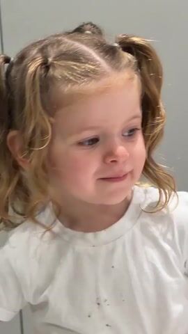 A young girl with blond hair in pigtails smiles at the camera. She is wearing a white T-shirt. She is indoors with a light-colored background. The child’s mouth opens slightly, then she grins. She turns her head to the left. Upbeat music plays in the background.