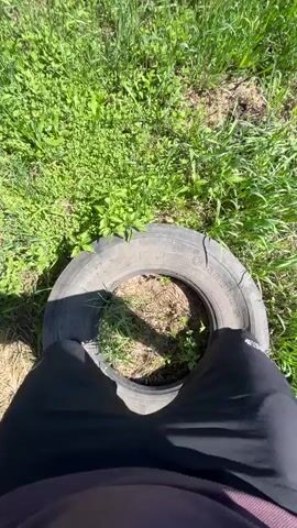 The video, filmed from a first-person perspective, shows a person performing an exercise outdoors. The person's legs, wearing black shorts or pants and black sneakers, are visible as they repeatedly step into and out of the center of a gray tire lying flat on the ground. The tire appears to be a car or small truck tire, and the text "CAROLUS" is partially visible on its side, along with other unreadable markings. The exercise involves quick, rhythmic steps over and through the tire. The setting is a grassy outdoor area, possibly a yard or field, with bright green grass and some other vegetation underfoot. The activity takes place on a sunny day, indicated by the clear shadows cast on the ground.