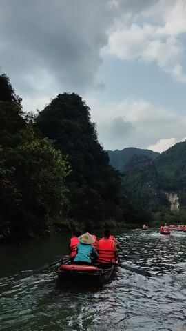 The video shows a group of people riding in rowboats on a river surrounded by lush, green mountains under a cloudy sky. The boats are filled with people wearing orange life vests, and some are also wearing traditional Vietnamese conical hats. The water is dark and choppy, reflecting the overcast sky. The mountains are densely covered in trees, creating a dark green backdrop against the lighter sky. The boats are moving forward, suggesting a guided tour or scenic ride through the river.