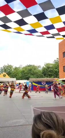 The video captures a vibrant outdoor cultural performance taking place during the daytime in what appears to be a paved courtyard or plaza. Overhead, a large, festive canopy composed of black, white, yellow, red, and blue checkered fabric squares provides shade.

The central activity features a group of approximately eight to ten performers, likely dancers, dressed in elaborate and highly colorful costumes. These costumes are adorned with numerous fringes, ribbons, and intricate patterns in bright hues of red, yellow, green, and blue, suggesting traditional attire. Several performers also wear what appear to be mask-like elements or large, plumed headdresses. They are engaged in an energetic and synchronized dance across the concrete ground, with fluid movements and raised arms.

In the background, a large Venezuelan flag or banner is prominently displayed on a stage-like structure, indicating the location is Venezuela, specifically Cabudare as per the context. Behind this, lush green trees and foliage line the horizon. To the right of the frame, a modern, multi-story building constructed with red brick features several dark, rectangular windows. A person's head and hair are partially visible in the bottom foreground, suggesting the presence of spectators. The scene conveys a festive and celebratory atmosphere.