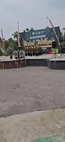 A group of children in matching brown uniforms, likely Muslim students, are standing in a line on a raised stage. They are performing a synchronized hand gesture, possibly a religious song or dance. Behind them is a large black banner with text that reads "FUN CAMP AKBAR #2" and "Hizbul Wathon," along with some logos and decorations. Two empty white chairs are placed in front of the line of children. To the left of the stage, two individuals in similar brown uniforms stand apart from the main group, facing the audience. The stage is made of concrete and has a dark-colored border. In front of the stage is a large, open concrete area. The background shows trees and a cloudy sky, suggesting an outdoor event. There are also flags, a speaker on a stand, and some equipment that appears to be part of a playground or amusement ride in the distance.